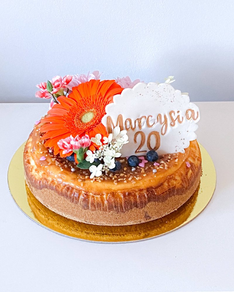 Sernik udekorowany pomarańczową gerberą, drobnymi kwiatami, borówkami i topperem z napisem 'Marcysia 20'.