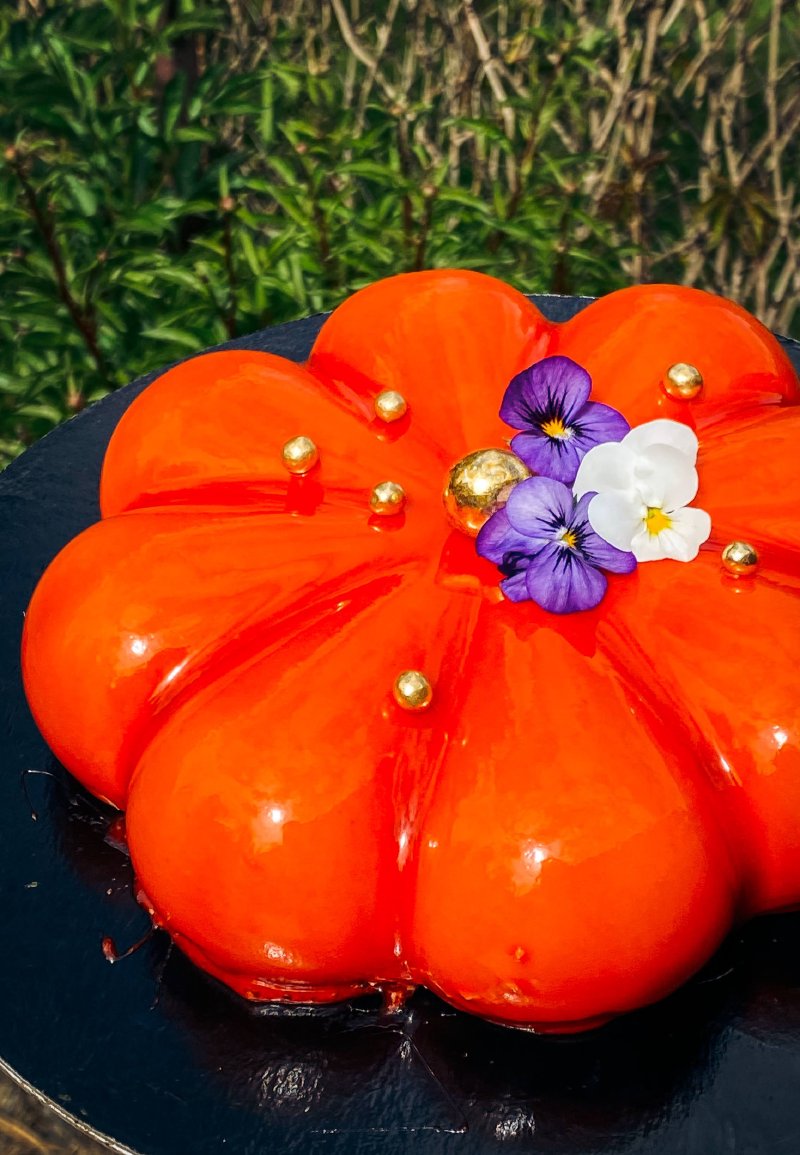 Pomarańczowy tort musowy w kształcie dyni z kwiatami fiołka i złotymi kuleczkami.
