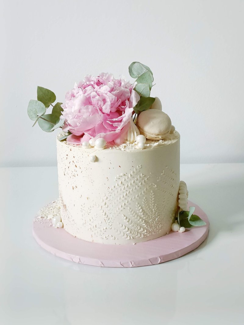 Elegancki biały tort z delikatnym zdobieniem i różową piwonią na wierzchu.