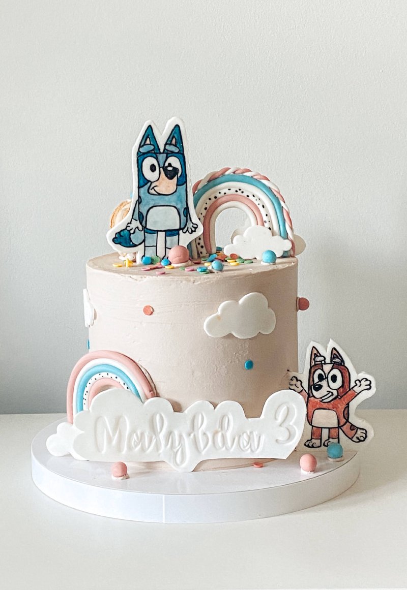 Pastelowy tort urodzinowy z postaciami Bluey i tęczowymi dekoracjami.