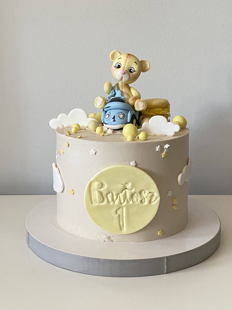 Okrągły, beżowy tort urodzinowy z figurką misia na samochodziku i personalizowanym emblematem.