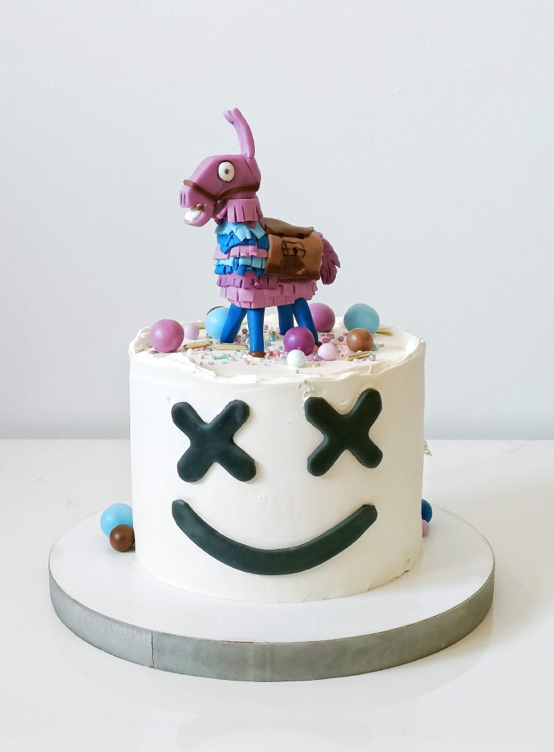 Biały tort urodzinowy z emoji Marshmello i figurką Llama Piñata na wierzchu.
