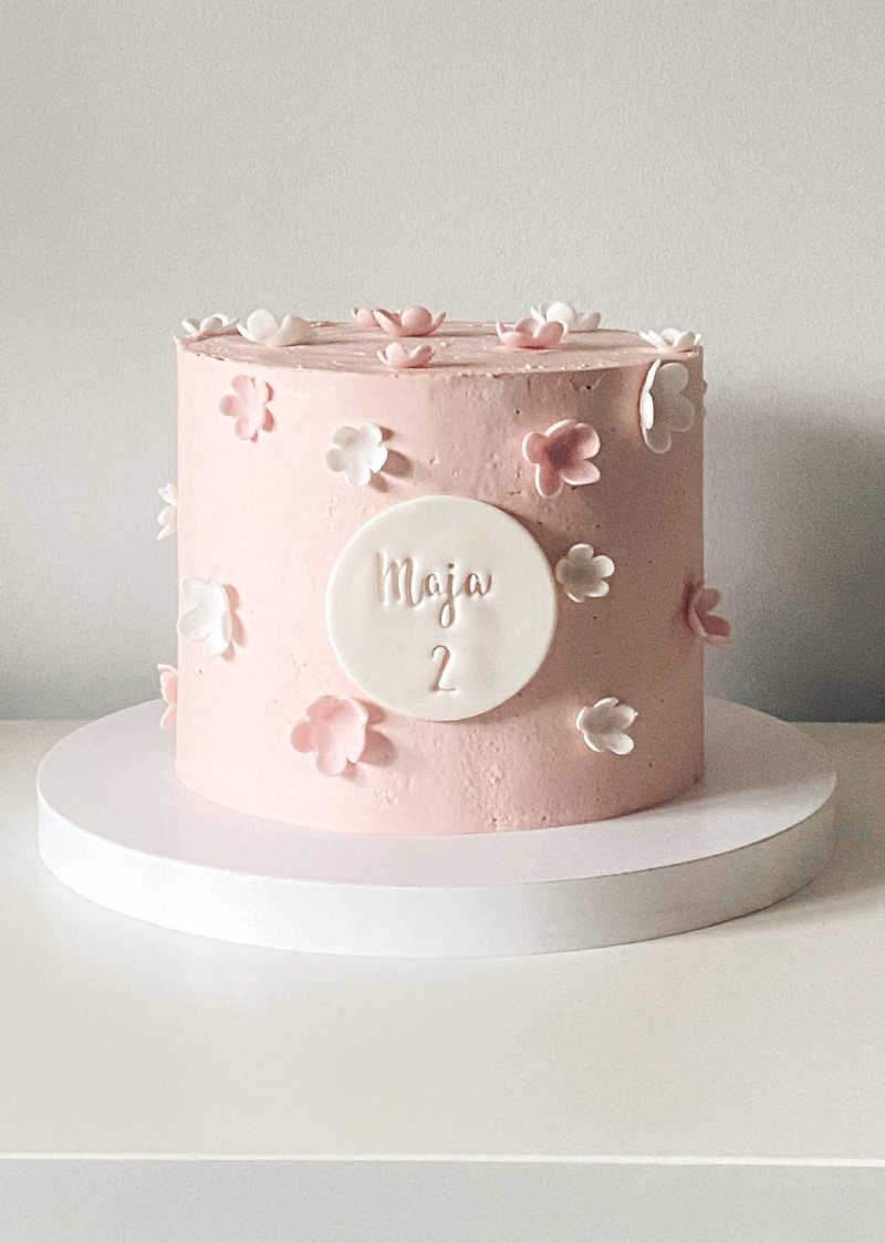 Różowy tort urodzinowy ozdobiony małymi, cukrowymi kwiatuszkami i personalizowaną plakietką.