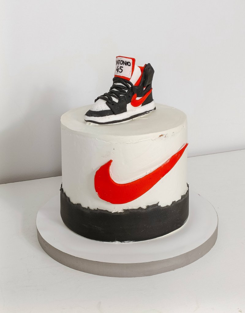 Biały tort urodzinowy z czerwonym logo Nike i figurką buta sportowego na wierzchu.
