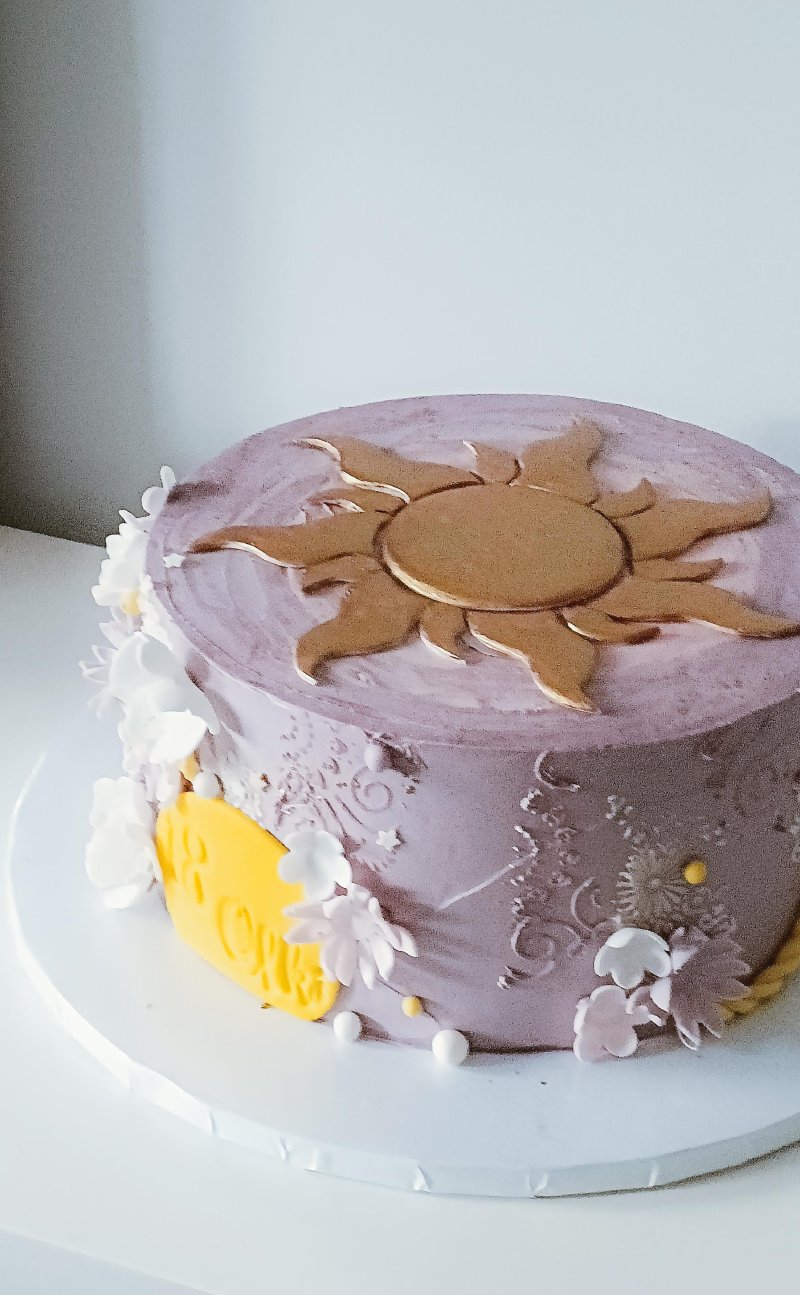 Elegancki tort urodzinowy w lawendowym kolorze ze złotym emblematem słońca.