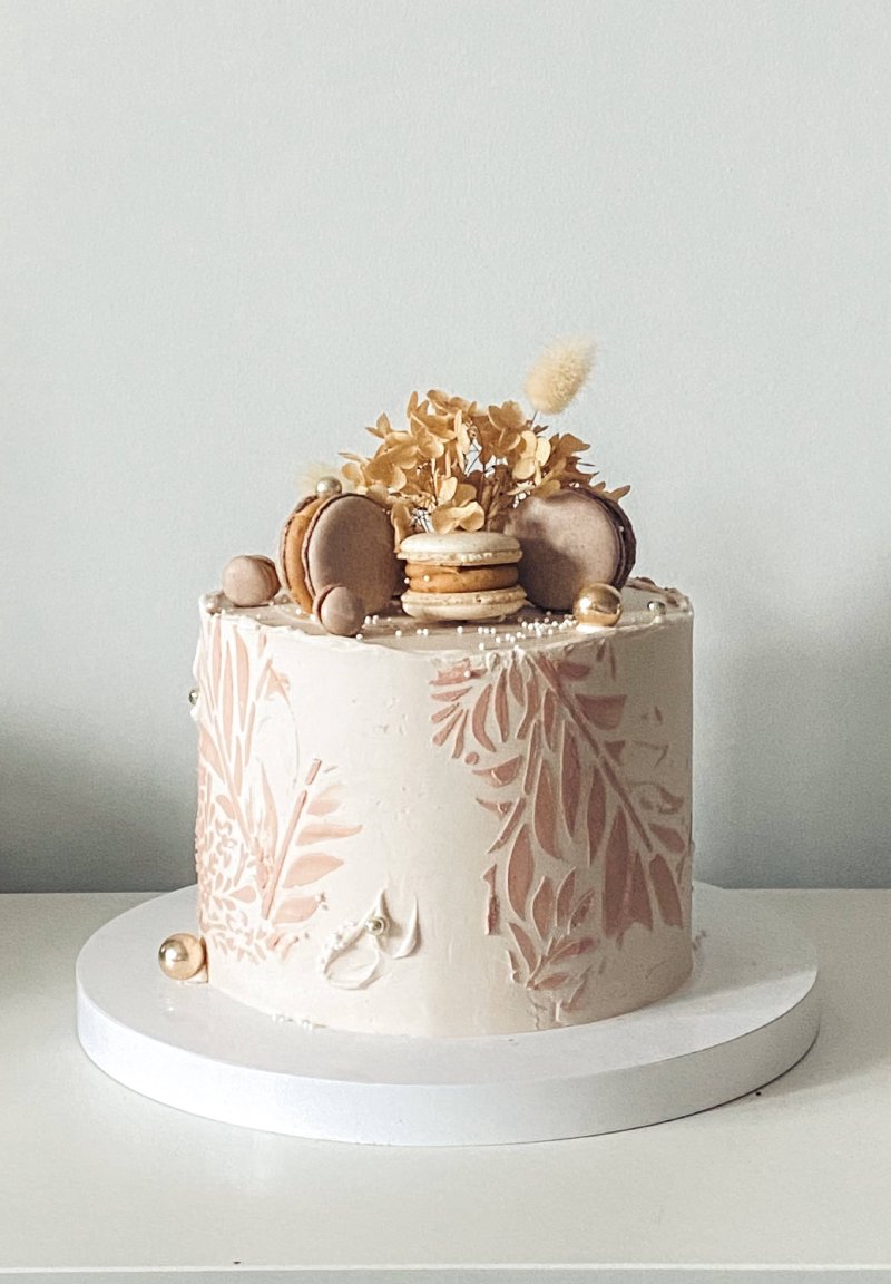 Elegancki tort z kremową polewą stemplowany wzorem botanicznym i udekorowany makaronikami.