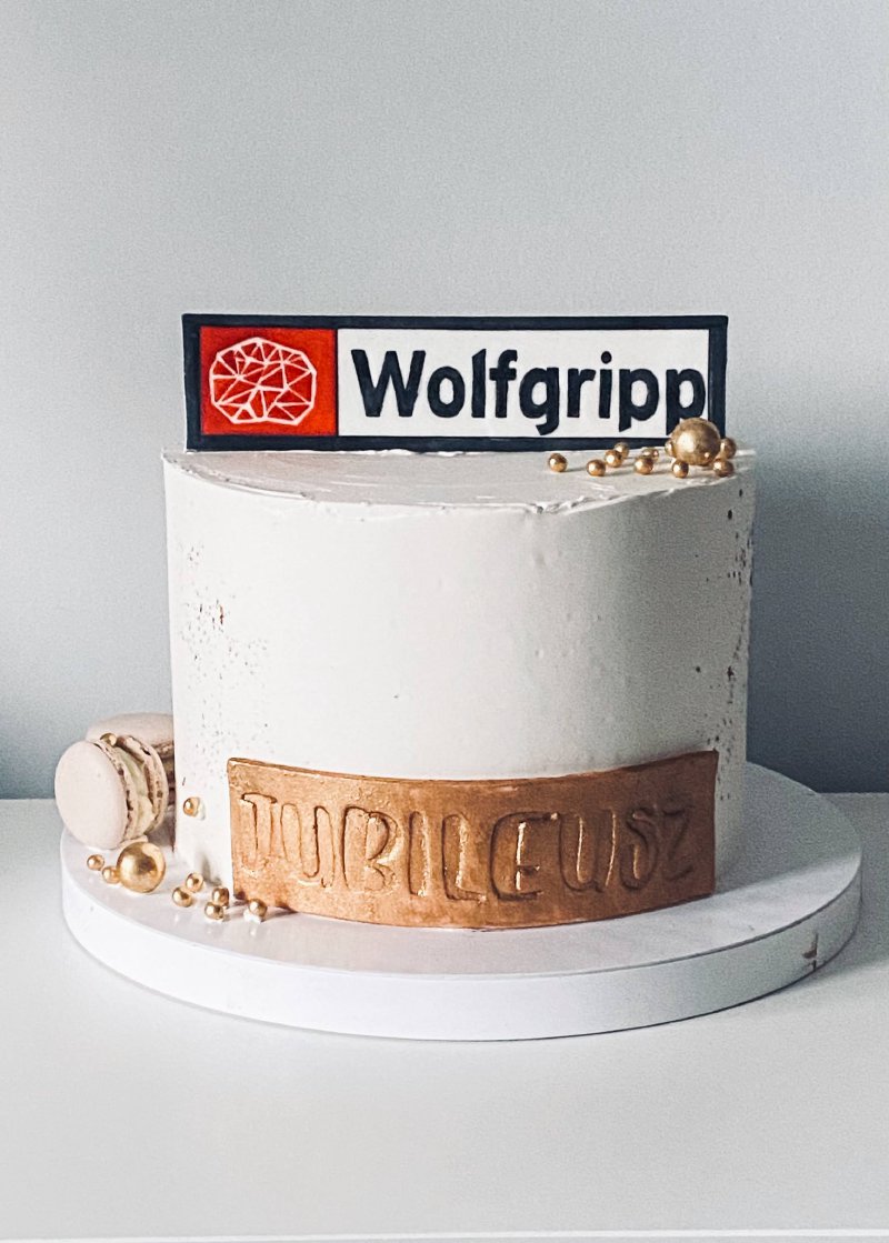 Biały tort z napisami 'JUBILEUSZ' i 'Wolfgripp' oraz złotymi akcentami