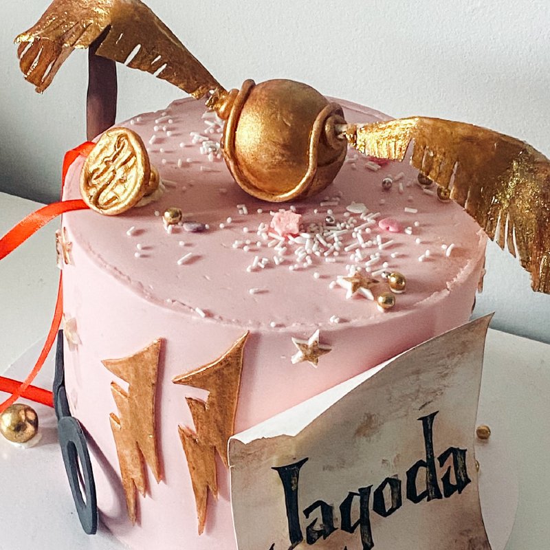 Różowy tort tematyczny ozdobiony złotym Zniczem i dekoracjami Harry'ego Pottera